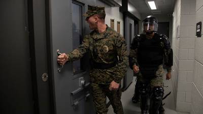A male Marine in camis in the center opens the door to a brig cell. Another male Marine in protective gear trails behind him.