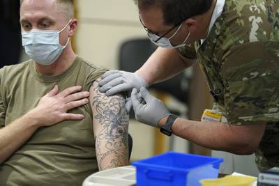 A seated and masked soldier receives a vaccination shot in his tattooed arm.