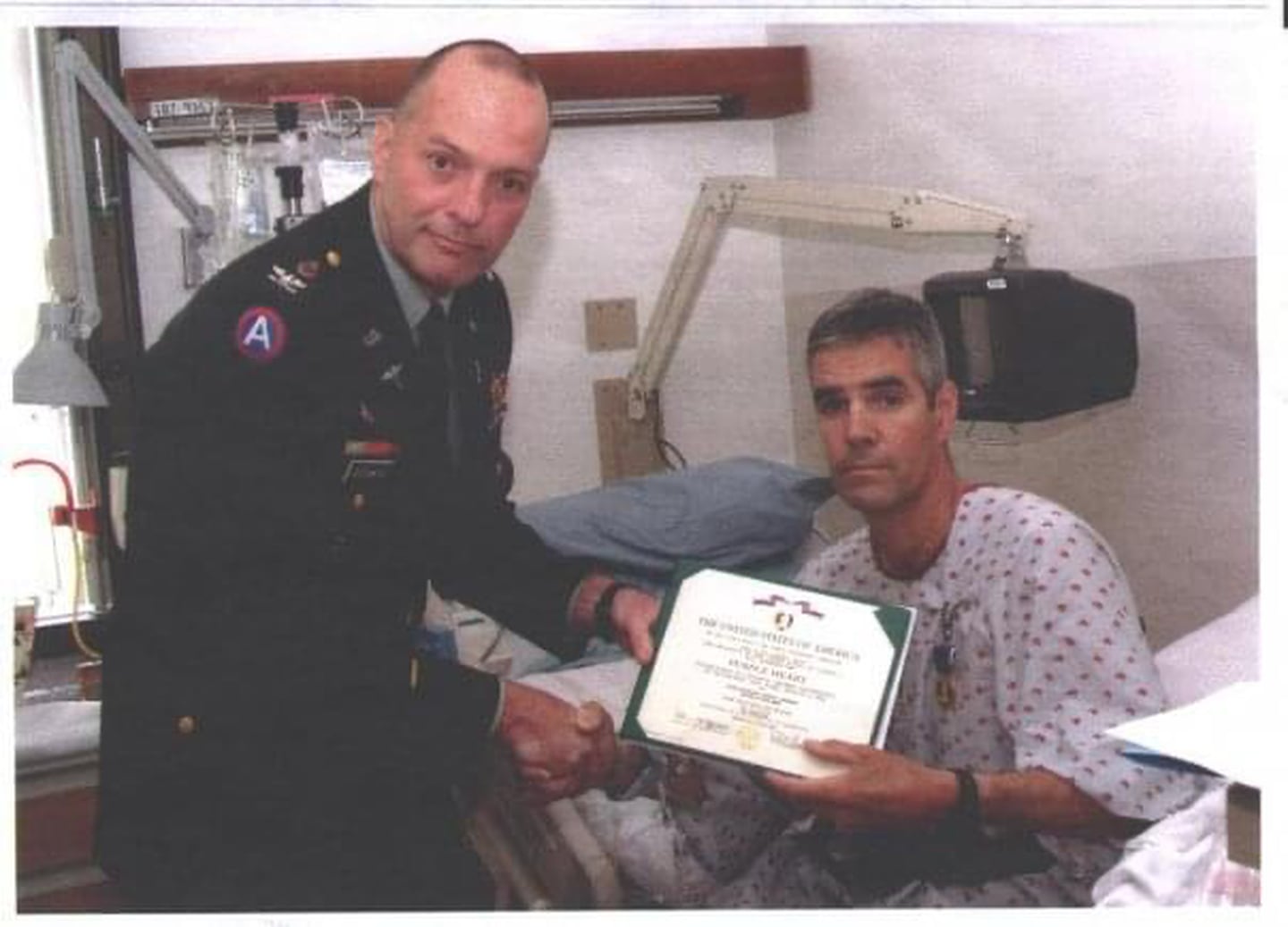 Army Sgt. 1st Class Joe Bowser sits in his hospital bed and holds a certificate for his Purple Heart award as he shakes the hand of the individual presenting him the award.