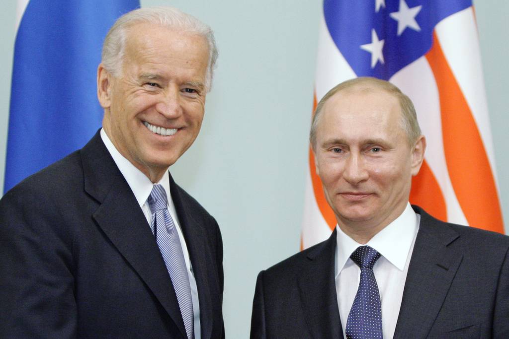 Joe Biden shakes hands with Vladimir Putin in front of flags.