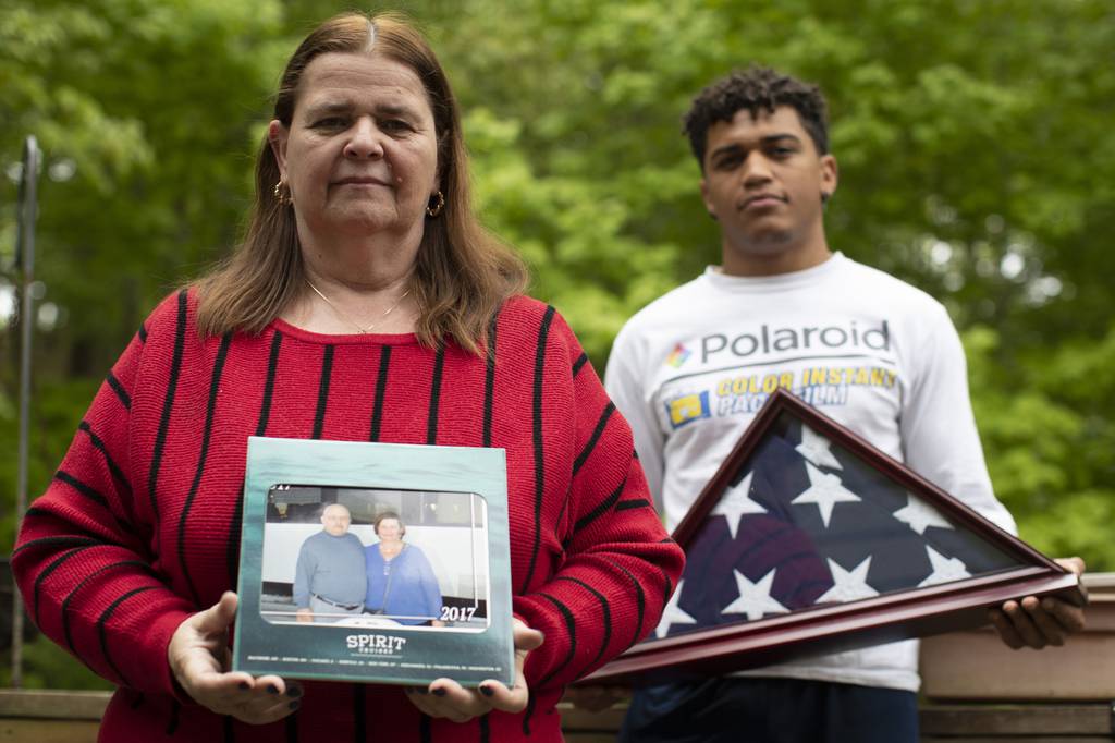 In this May 22, 2020, photo Florence Hopp, left, holds a photograph of herself and her husband Robert Hopp during a cruise in 2017, as her son J.J. Brania-Hopp holds the American flag the military presented to them after his father's death at their home in Boonton, N.J.